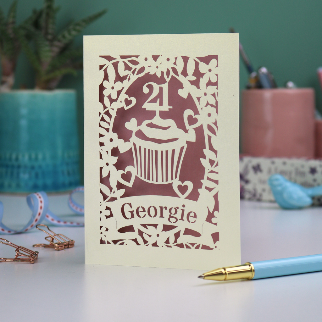 Decorative laser cut 21st birthday card with cupcake design and 'Georgie' text on a table with stationery items.