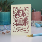 Decorative laser cut 21st birthday card with cupcake design and 'Georgie' text on a table with stationery items.