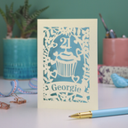 Decorative laser cut 21st birthday card with cupcake design and 'Georgie' text on a table with stationery items.