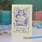 Decorative laser cut 21st birthday card with cupcake design and 'Georgie' text on a table with stationery items.