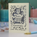 Decorative laser cut 21st birthday card with cupcake design and 'Georgie' text on a table with stationery items.
