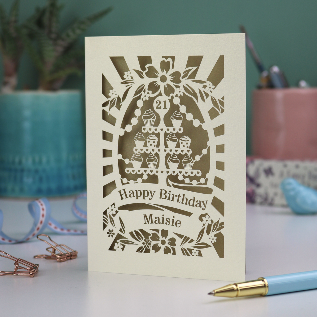 A paper cut personalised 21st Birthday card with decorative cupcake design and text on a desk with stationery items.