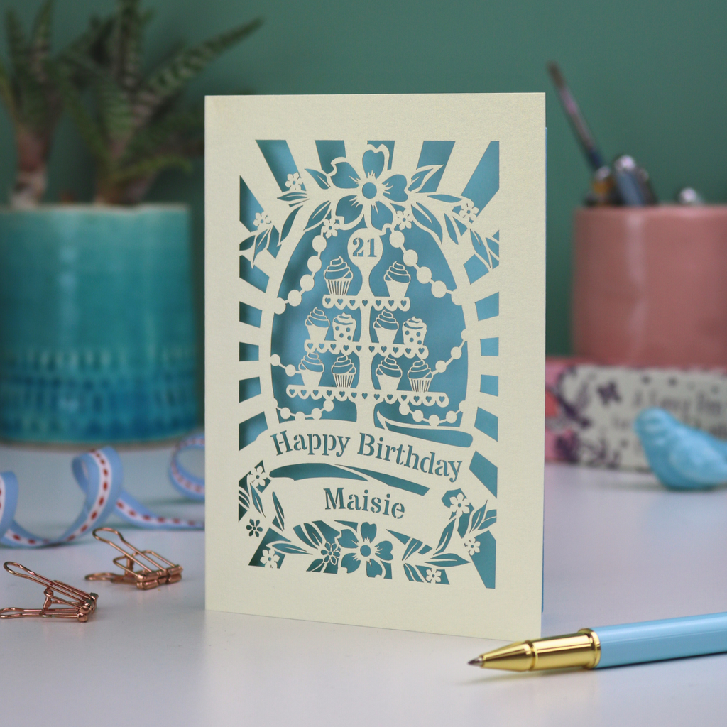 A laser cut personalised 21st Birthday card with decorative cupcake design and text on a desk with stationery items.