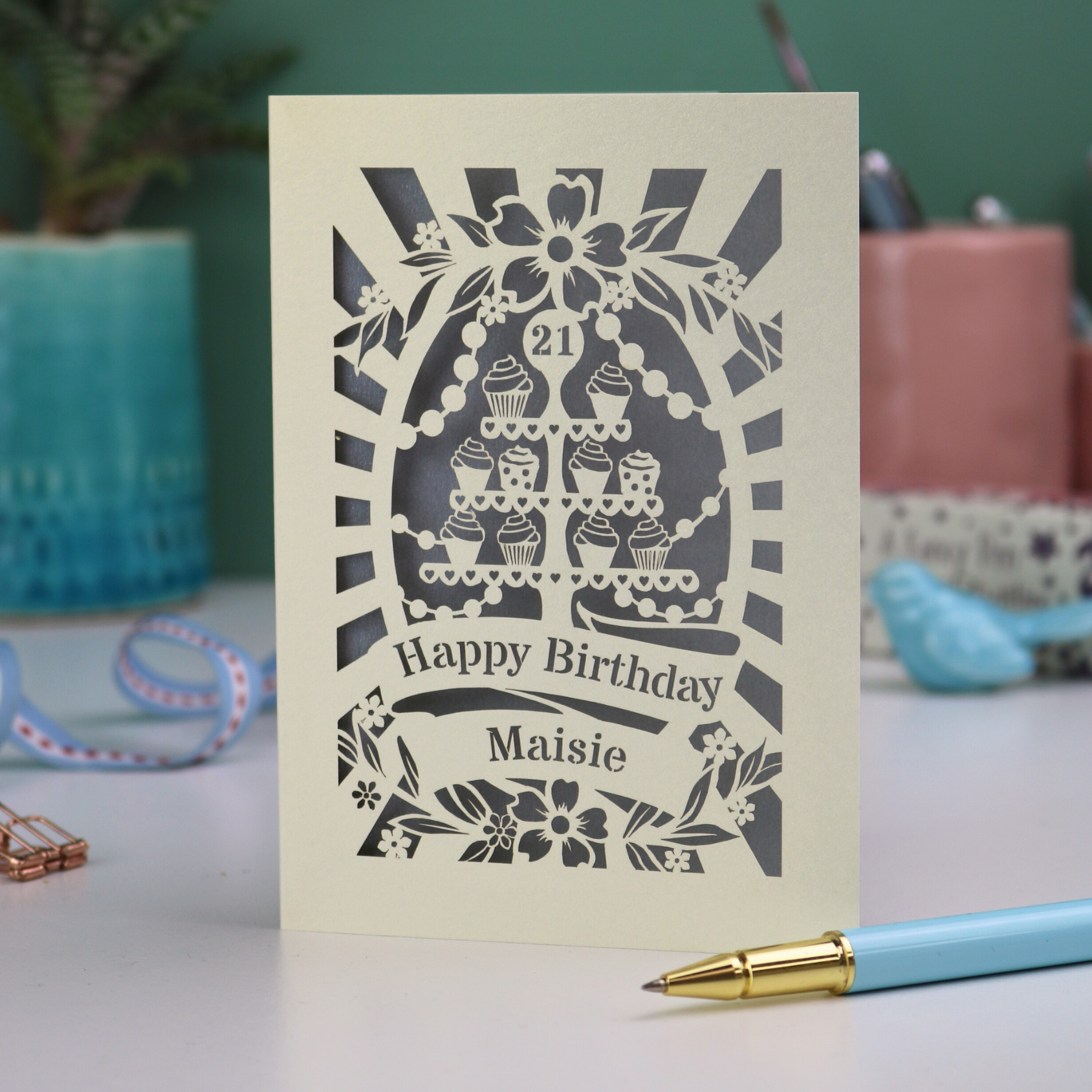 A laser cut personalised 21st Birthday card with decorative cupcake design and text on a desk with stationery items.