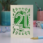 A laser cut personalised 21st birthday card with cut out spotty number 21 and name 'Robyn' on a table with a pen and decorative items.