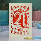 A paper cut personalised 21st birthday card with cut out spotty number 21 and name 'Robyn' on a table with a pen and decorative items.