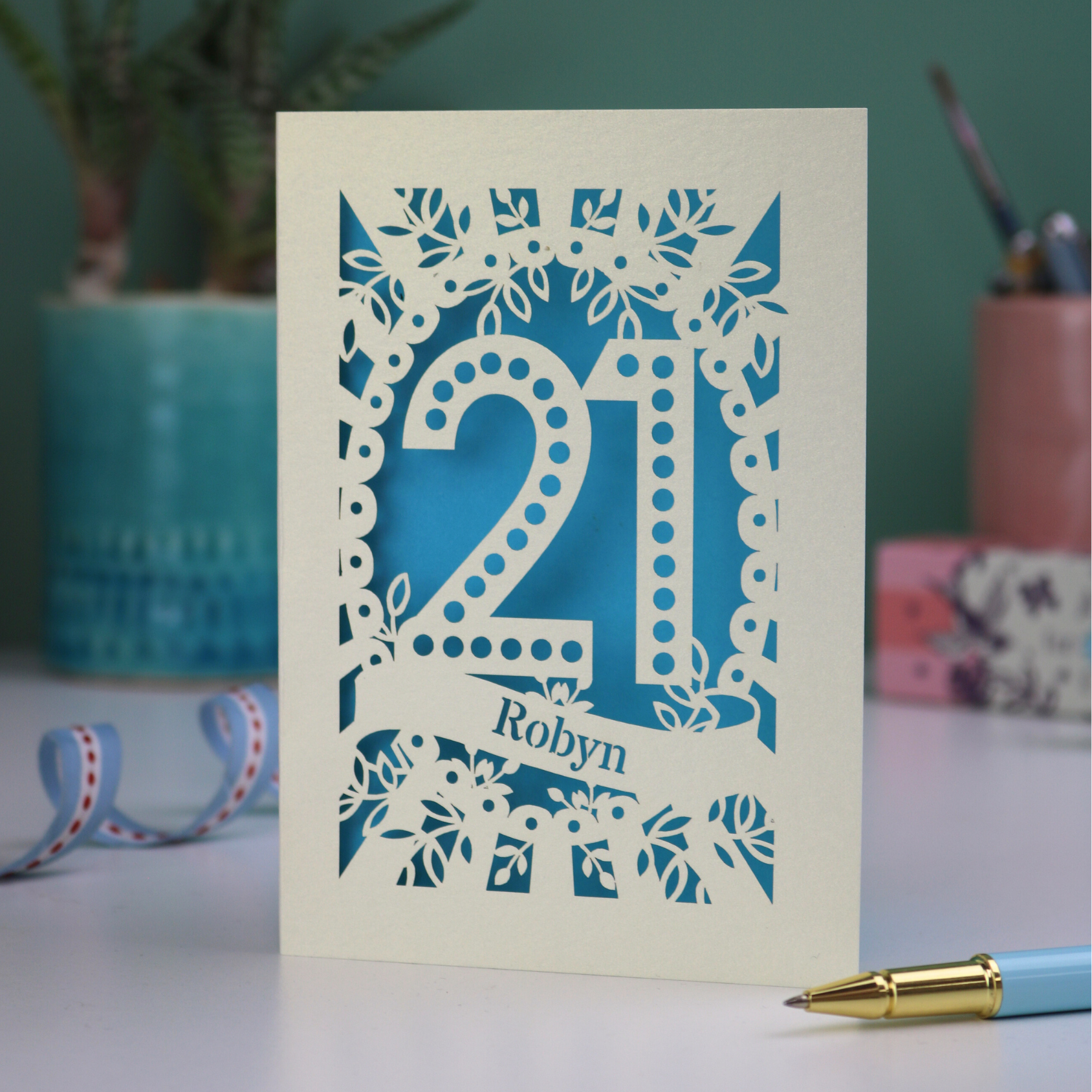 A laser cut personalised 21st birthday card with cut out spotty number 21 and name 'Robyn' on a table with a pen and decorative items.