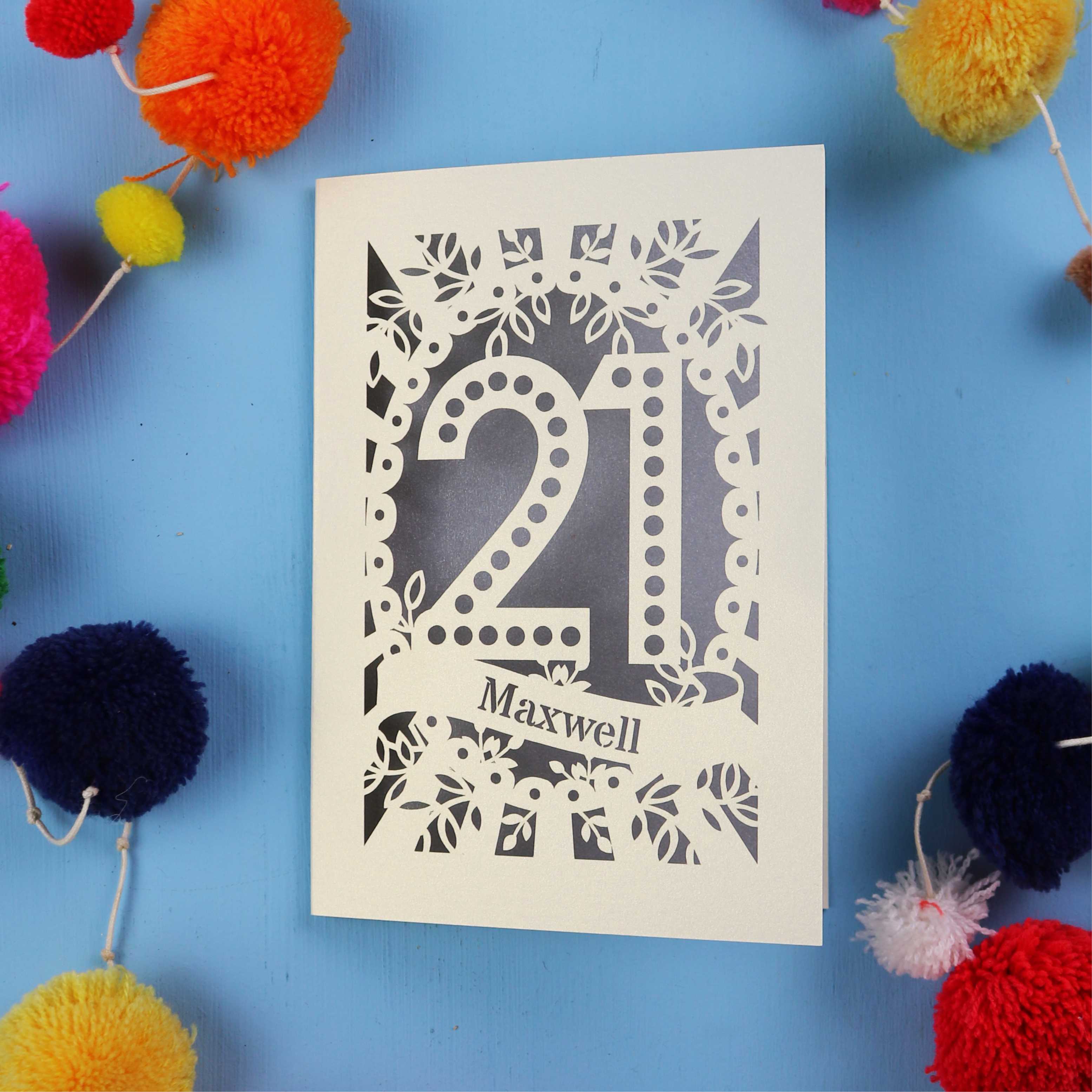 A 21st birthday Card with '21 Maxwell' design on a blue background with colourful pom-poms.