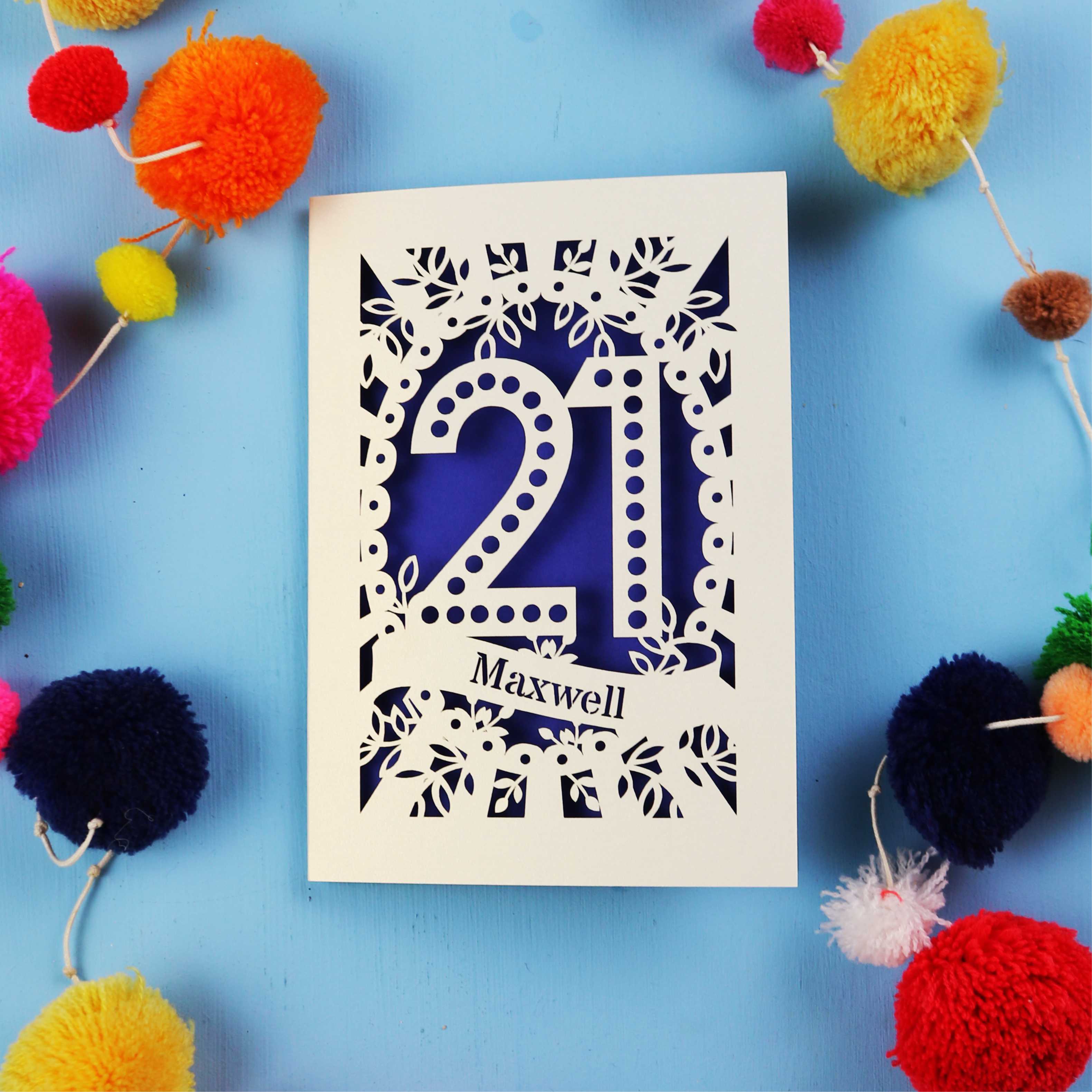 A 21st birthday Card with '21 Maxwell' design on a blue background with colourful pom-poms.