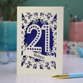 A laser cut personalised 21st birthday card with cut out spotty number 21 and name 'Robyn' on a table with a pen and decorative items.