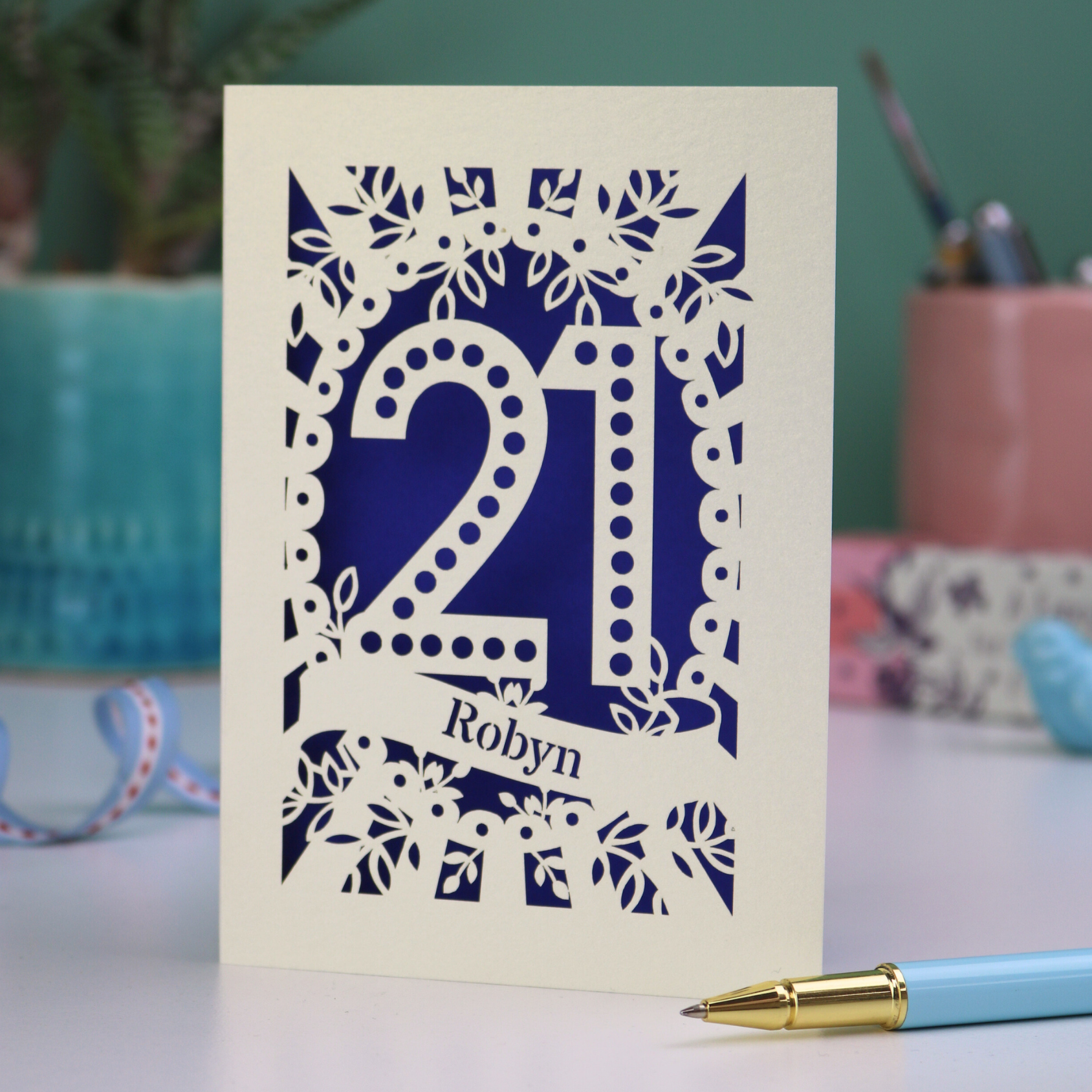A laser cut personalised 21st birthday card with cut out spotty number 21 and name 'Robyn' on a table with a pen and decorative items.