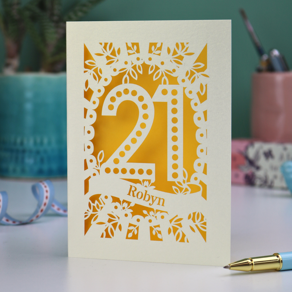 A keepsake personalised 21st birthday card with cut out spotty number 21 and name 'Robyn' on a table with a pen and decorative items.