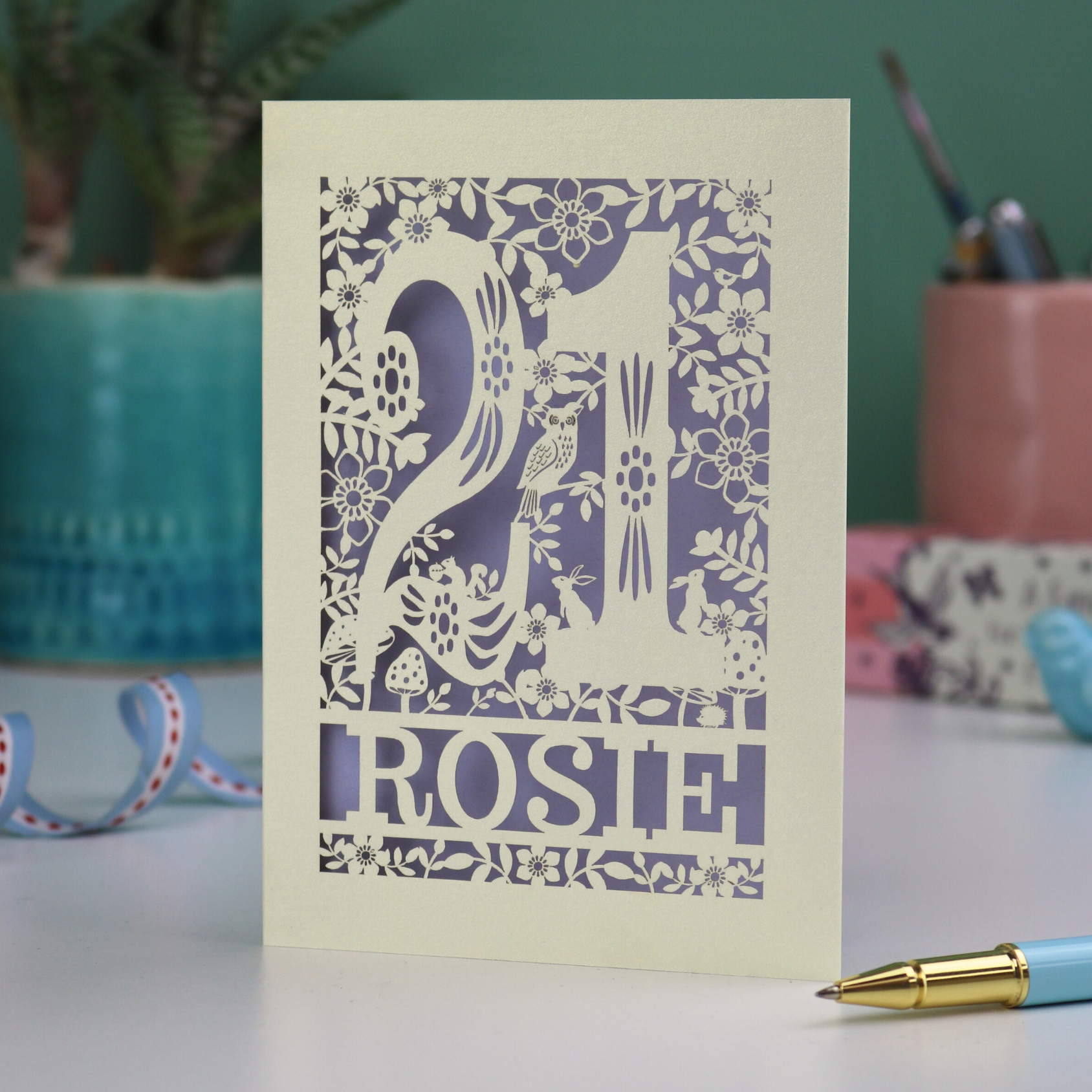 A laser cut 21st birthday card, personalised with the name "Rosie", with the number 21 in the centre and woodland theme design, including tiny rabbits, a squirrel, owl, toadstools, leaves and flowers.