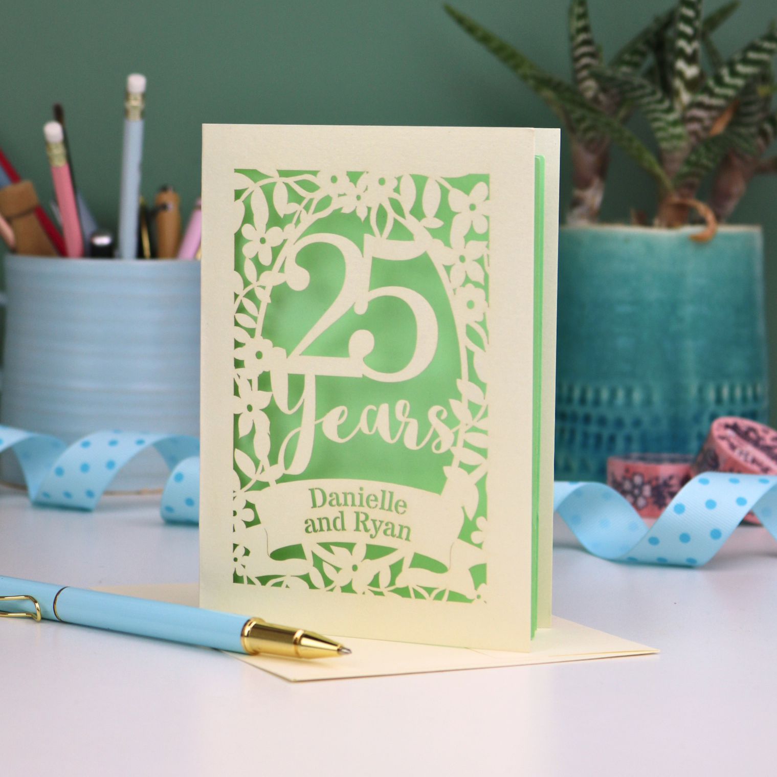 Silver anniversary Card with '25 Years' and 'Danielle and Ryan' on a desk with stationery items.