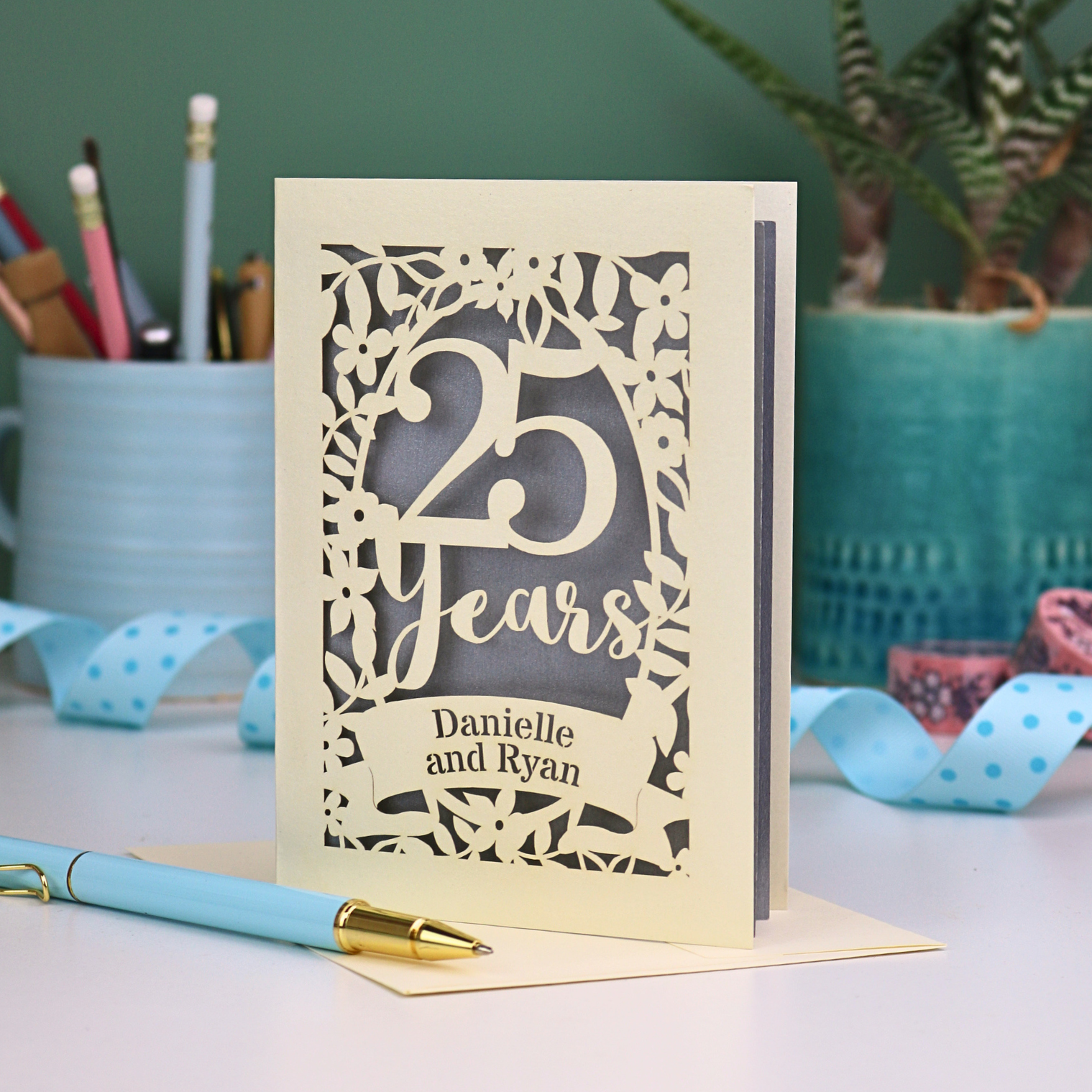 Silver anniversary Card with '25 Years' and 'Danielle and Ryan' on a desk with stationery items.