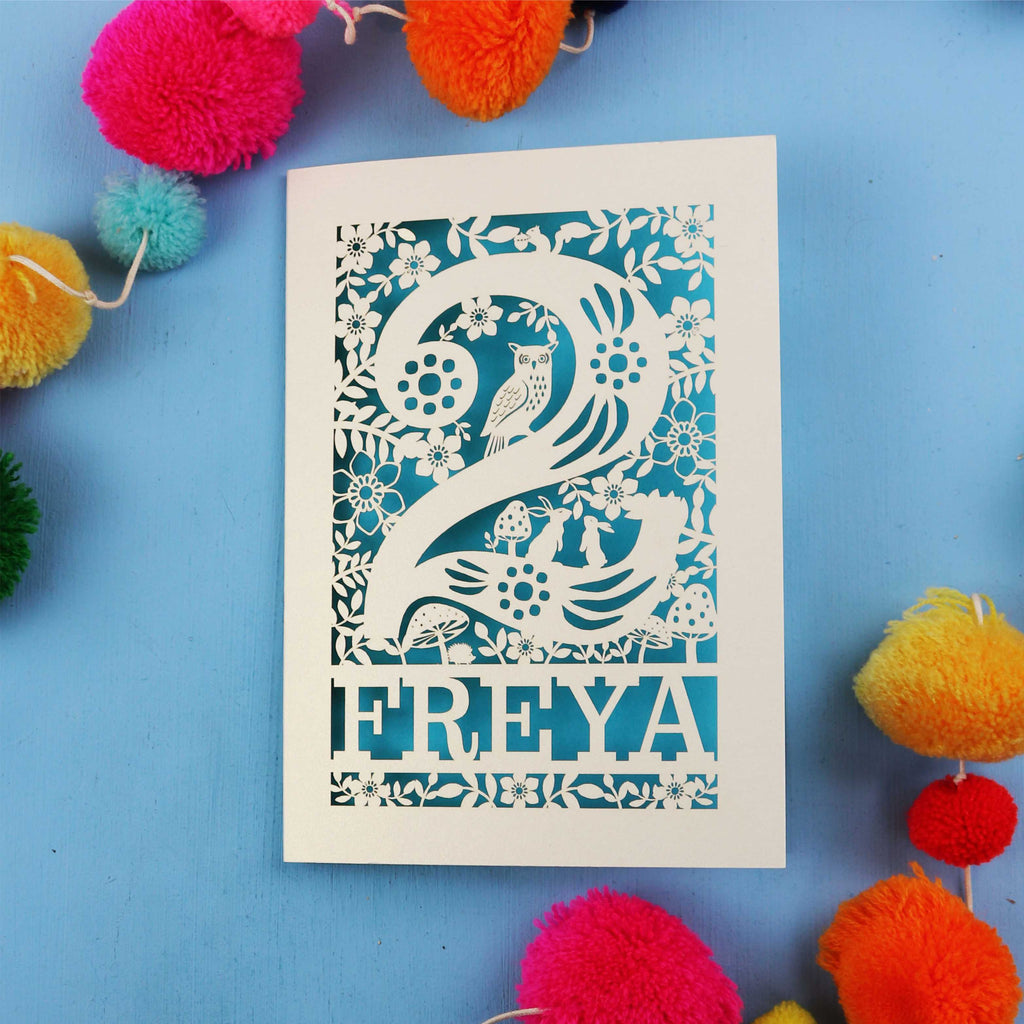 Decorative second birthday card with 'FREYA' design on a blue background with colourful pom-poms.