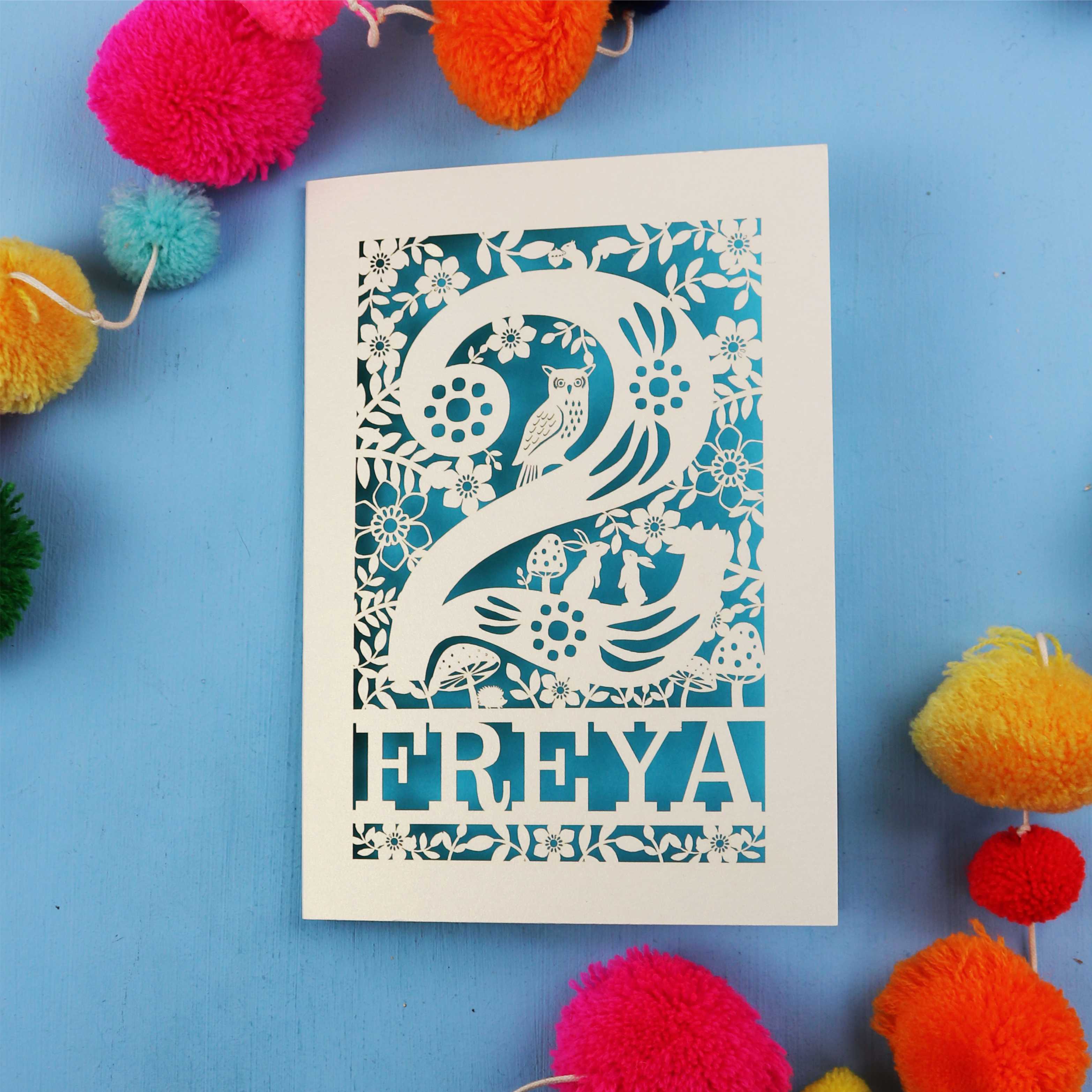 Decorative second birthday card with 'FREYA' design on a blue background with colourful pom-poms.