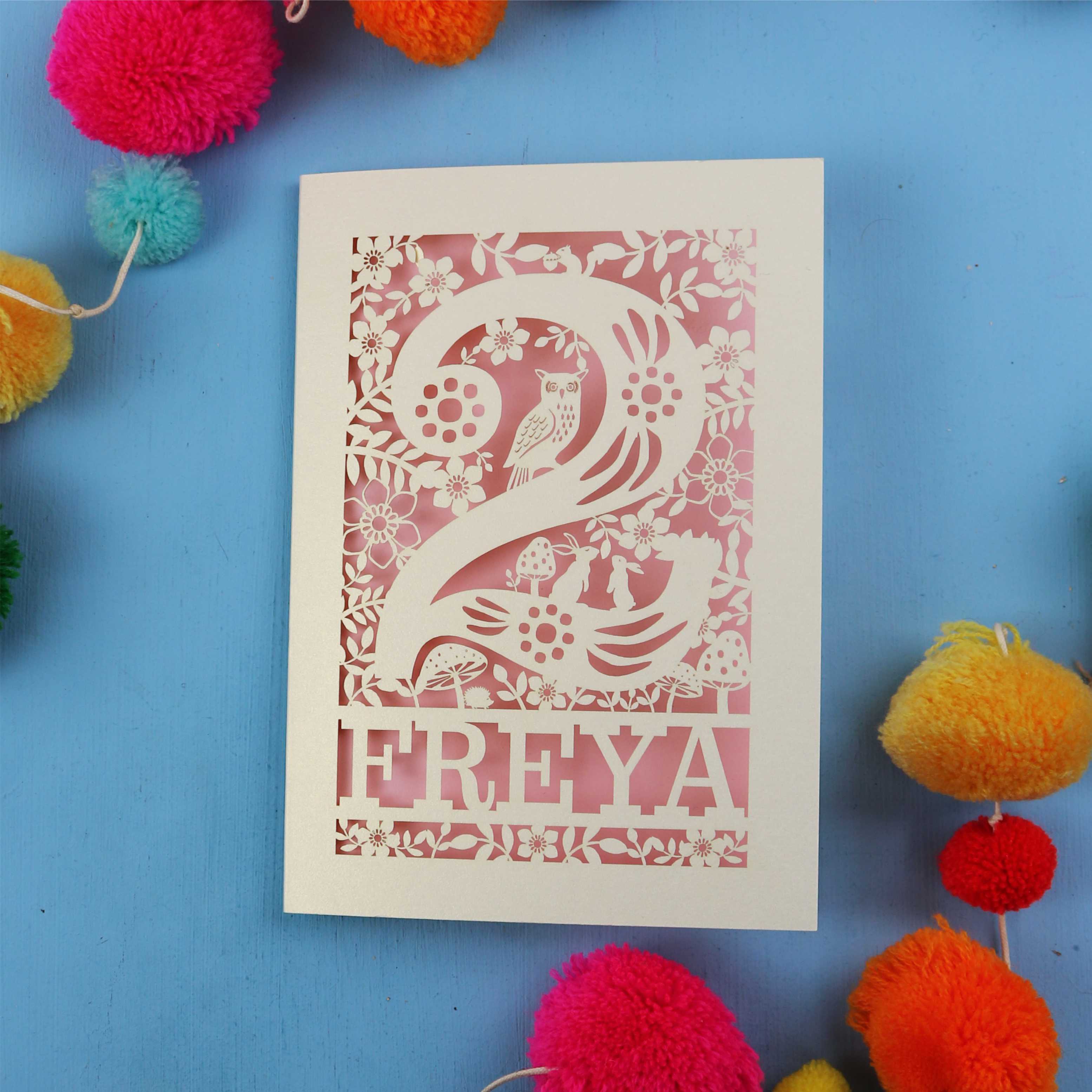 Laser cut 2nd birthday card with 'FREYA' design on a blue background with colourful pom-poms.