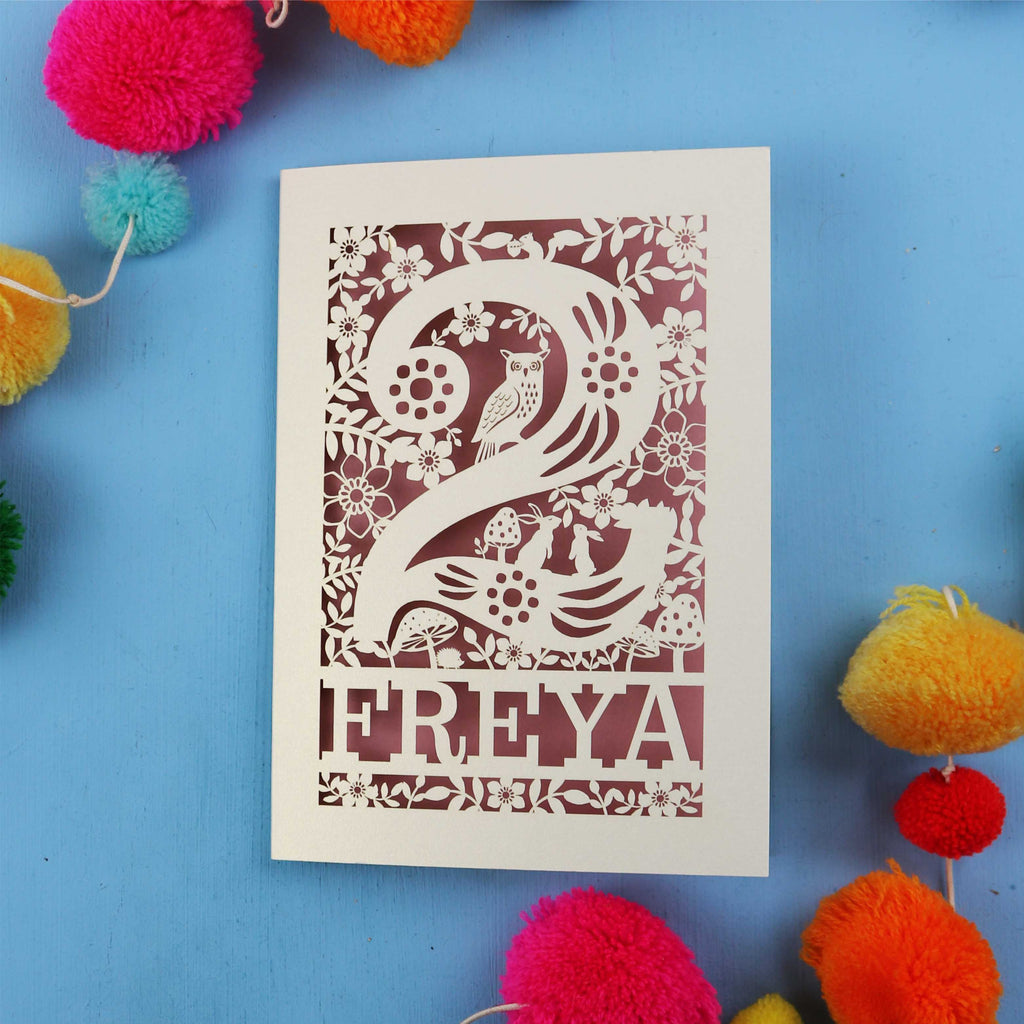 Laser cut second birthday card with 'FREYA' design on a blue background with colourful pom-poms.