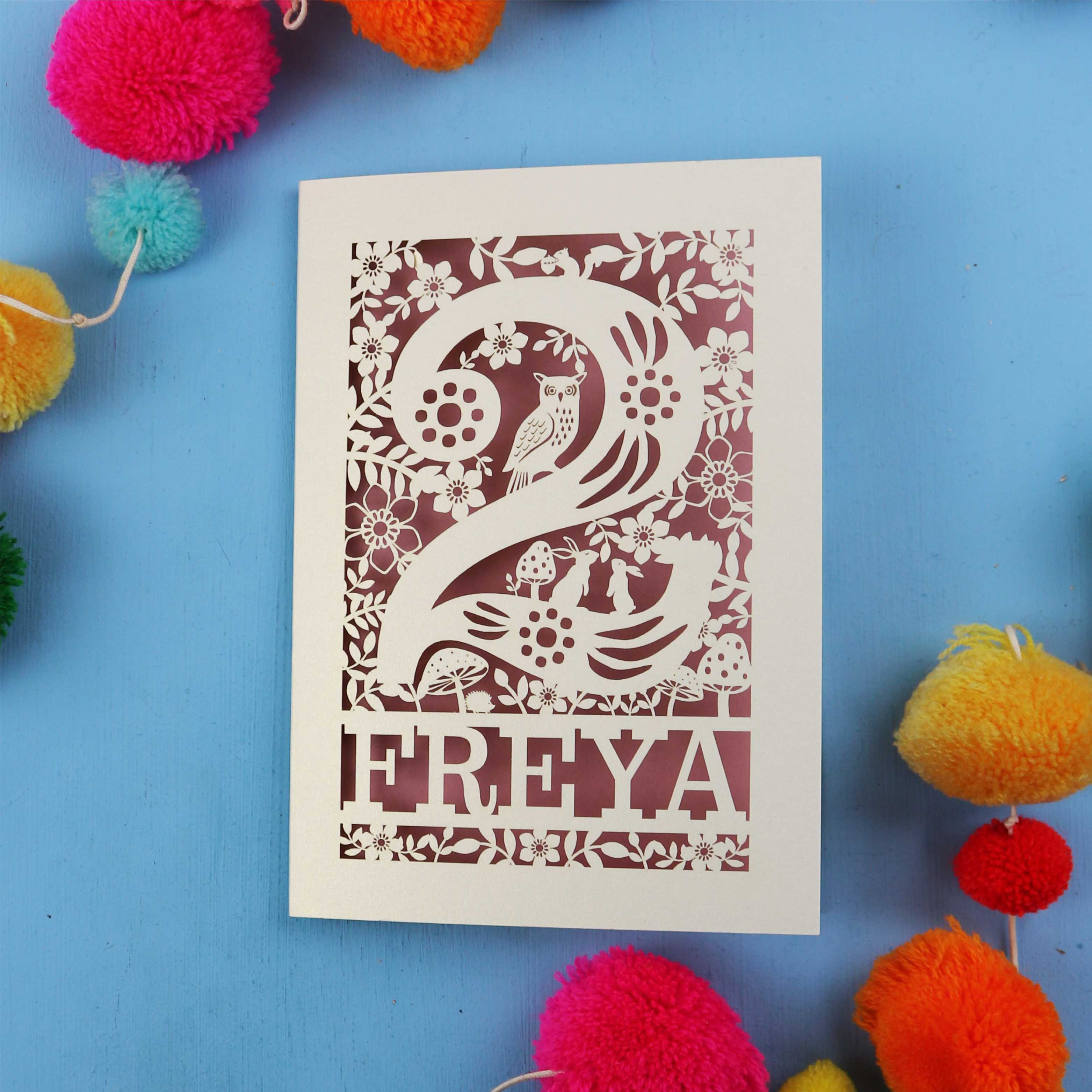 Laser cut second birthday card with 'FREYA' design on a blue background with colourful pom-poms.