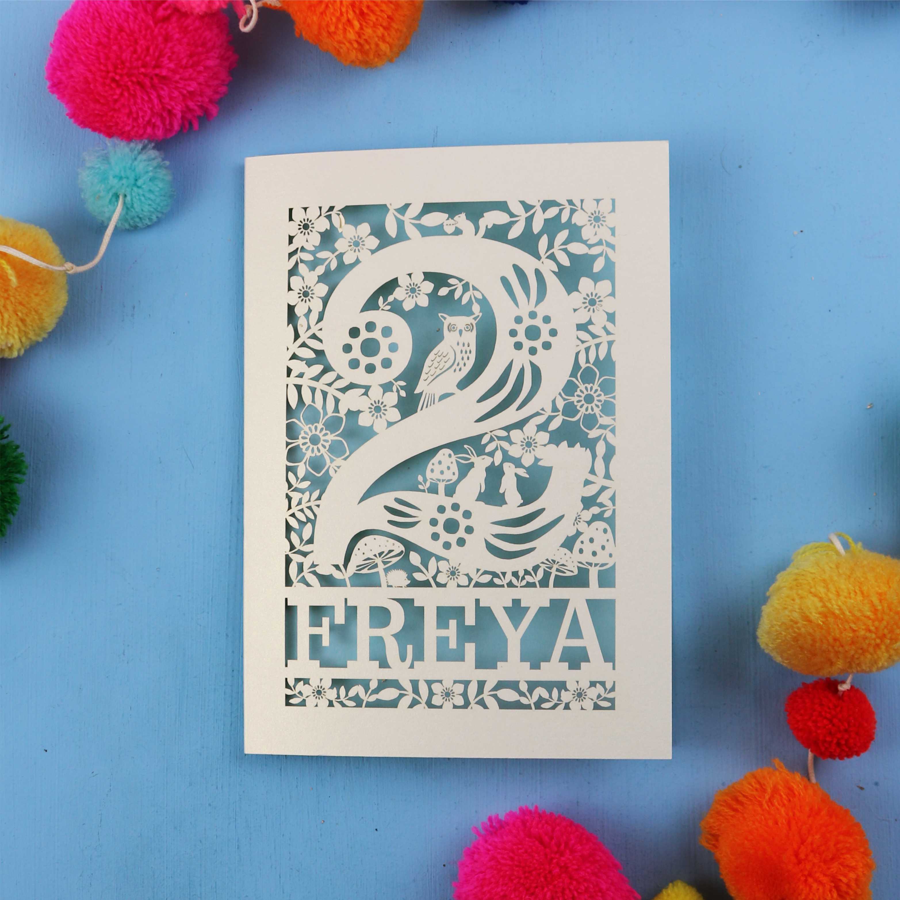 Laser cut 2nd birthday card with woodland design on a blue background with colourful pom-poms.