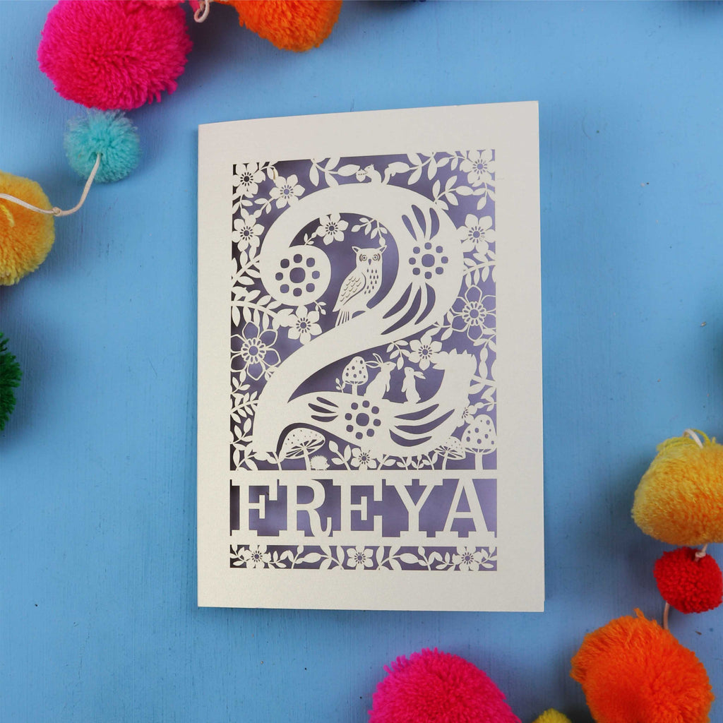 Laser cut second birthday card with woodland design on a blue background with colourful pom-poms.