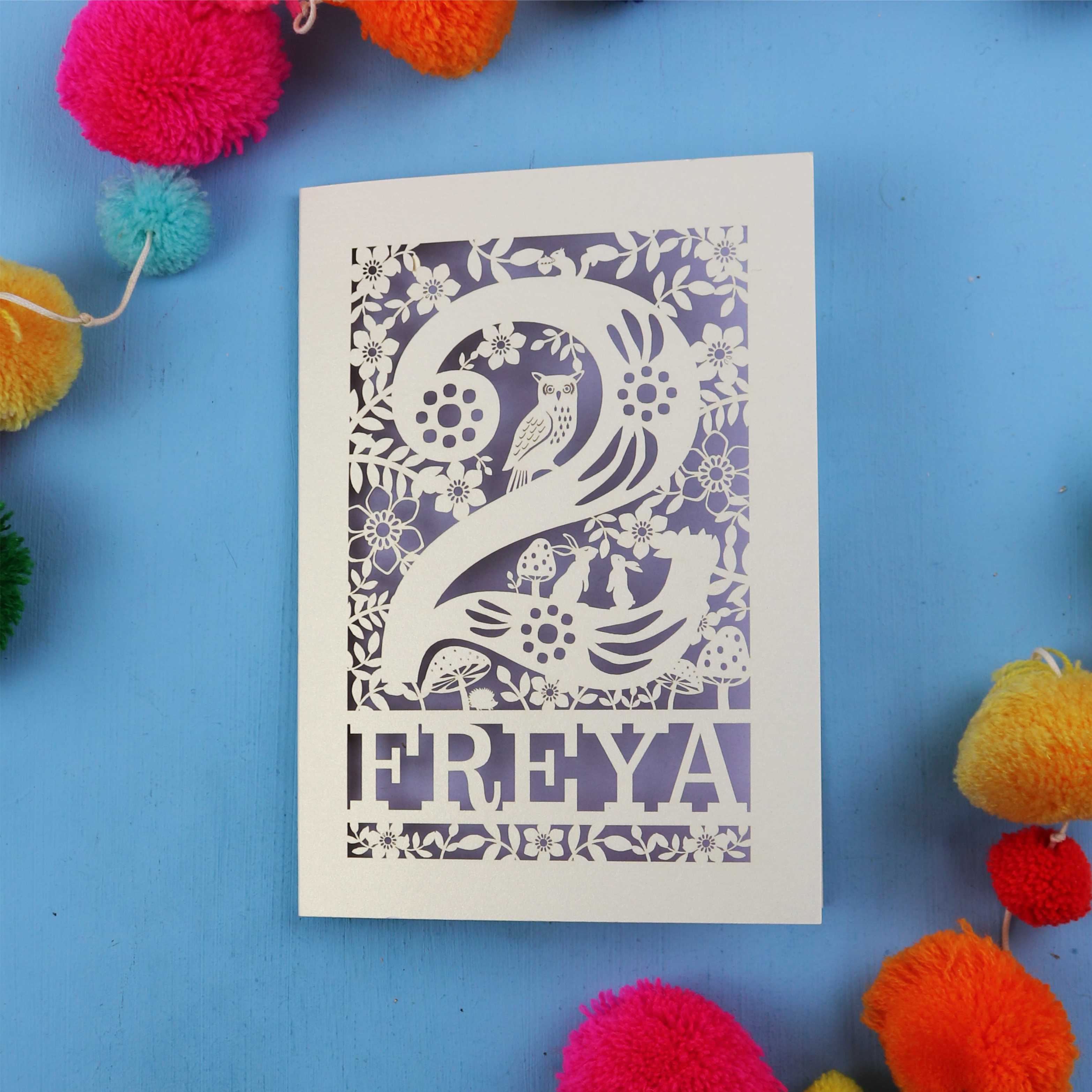 Laser cut second birthday card with woodland design on a blue background with colourful pom-poms.