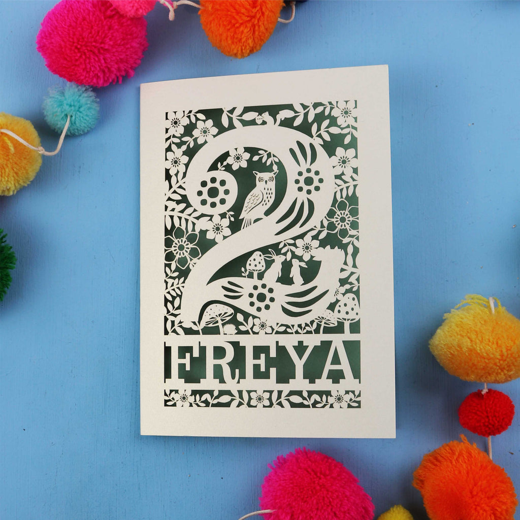Paper cut 2nd birthday card with woodland design on a blue background with colourful pom-poms.