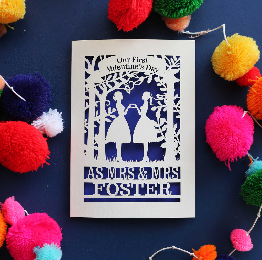 A personalised Valentine's day card with a laser cut design of two women making a heart shape, with a blank space for a personal message, and brightly coloured pom-pom decorations around it.