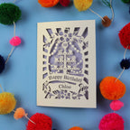 Decorative birthday card with cut-out design on a blue background with colorful pom-poms.