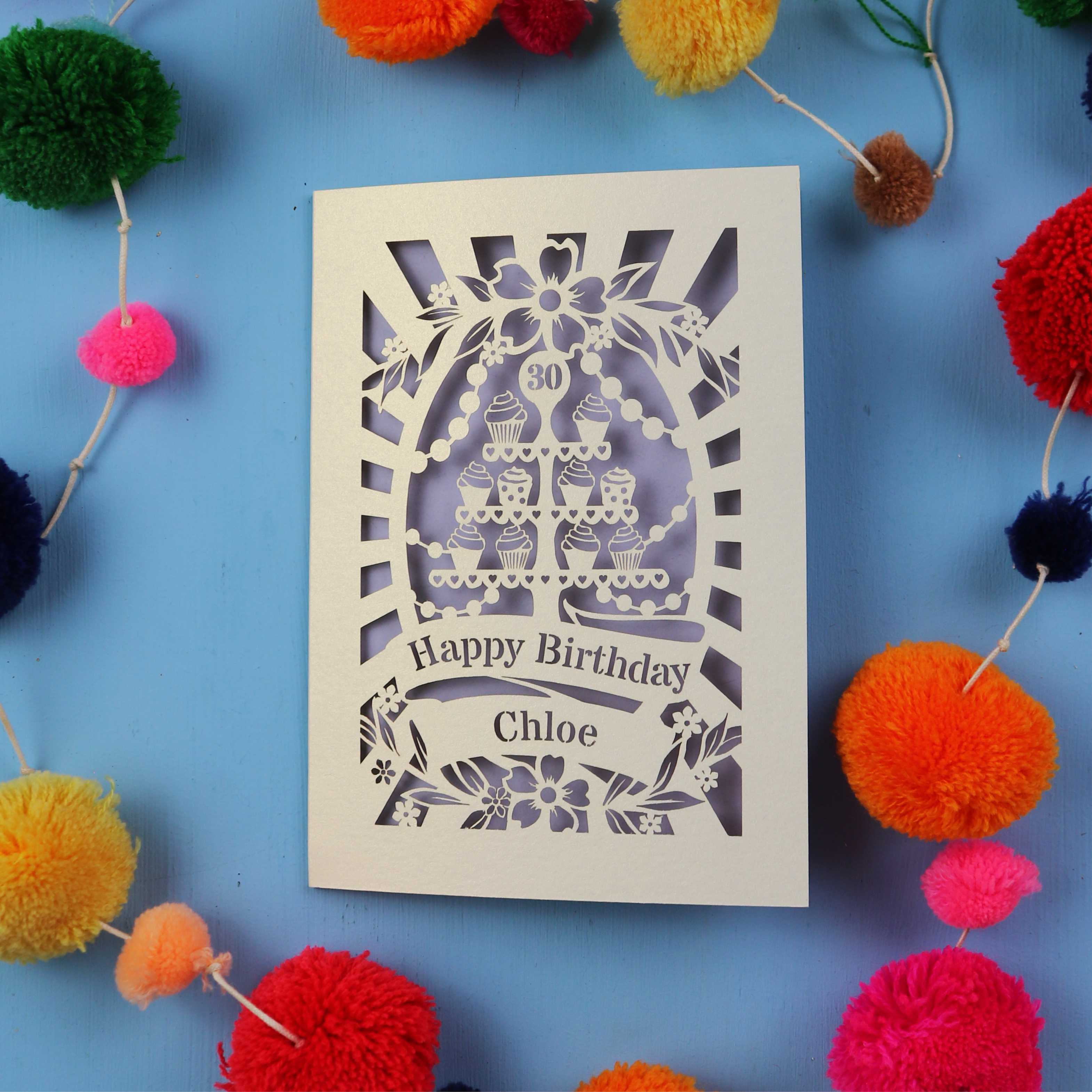 Decorative birthday card with cut-out design on a blue background with colorful pom-poms.