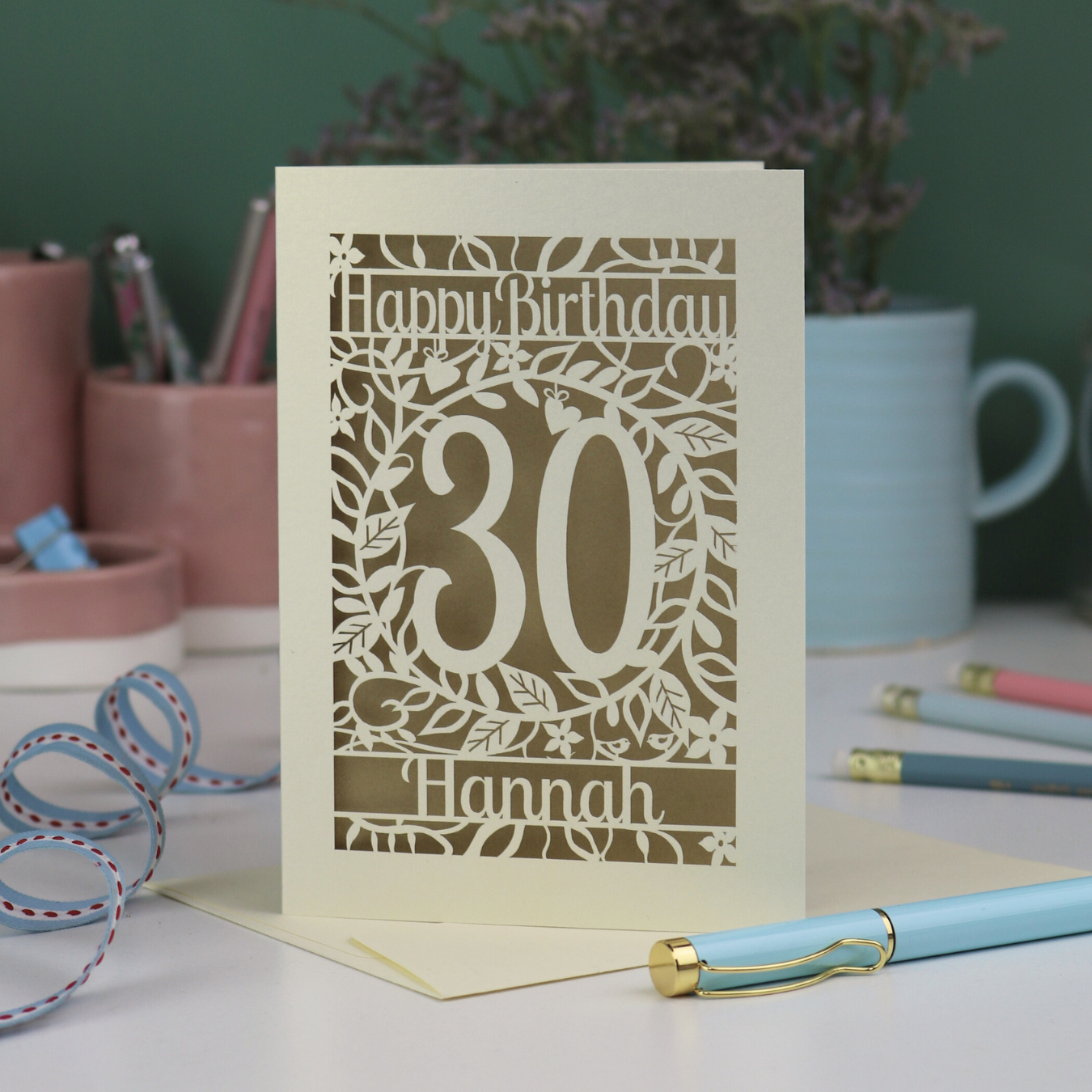 A laser cut floral 30th birthday card, personalised with the number 30 and the name Hannah, on a white desk with stationery items. 