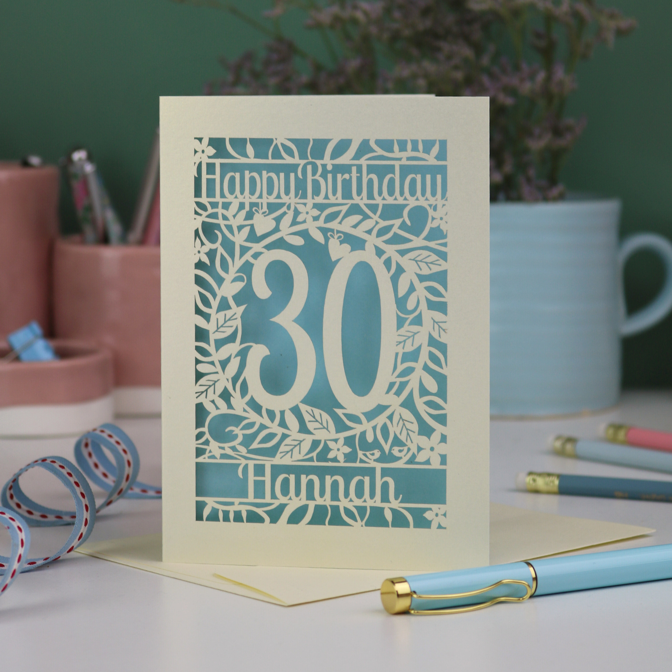 A cut out floral 30th birthday card, personalised with the number 30 and the name Hannah, on a white desk with stationery items. 