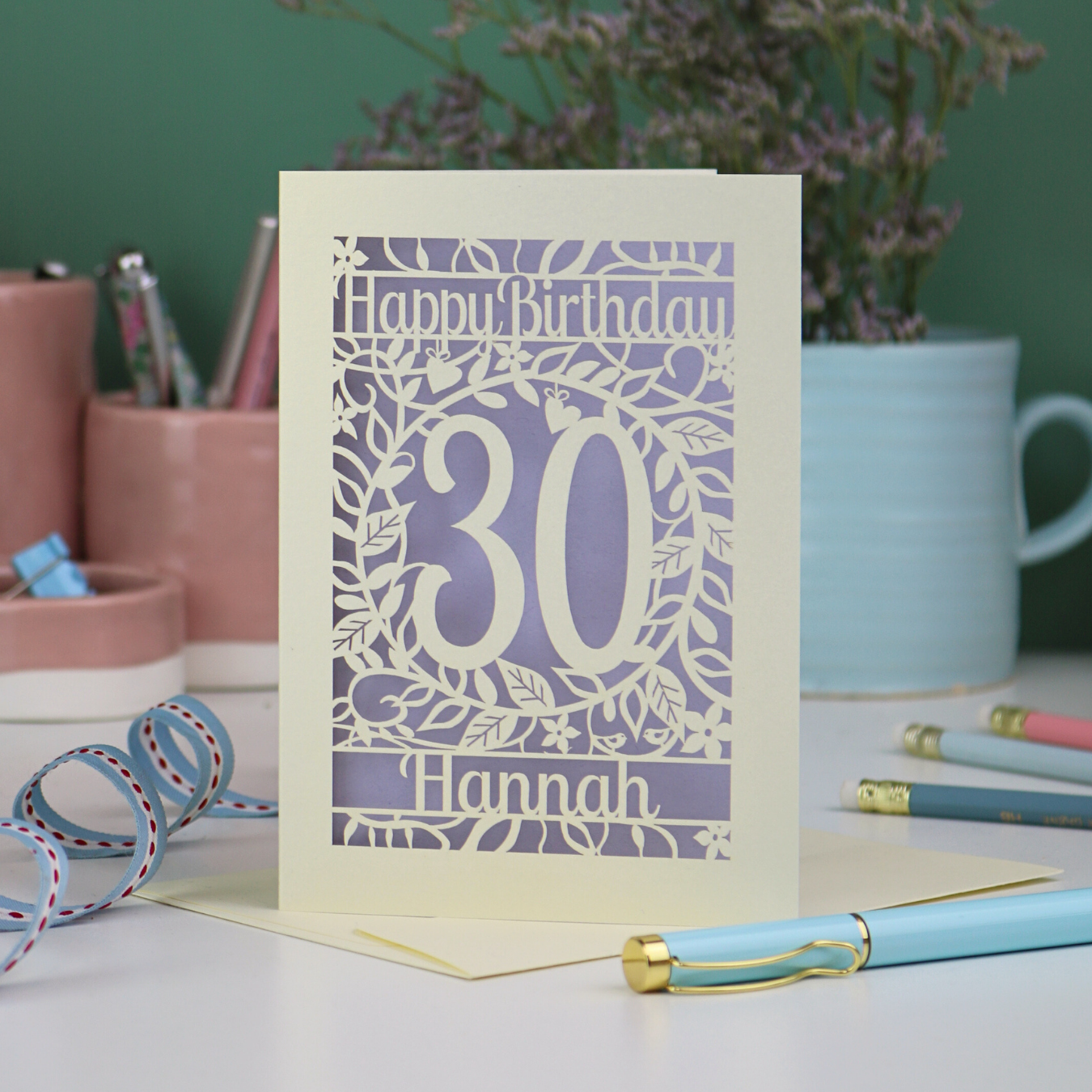 A laser cut floral 30th birthday card, personalised with the number 30 and the name Hannah, on a white desk with stationery items. 
