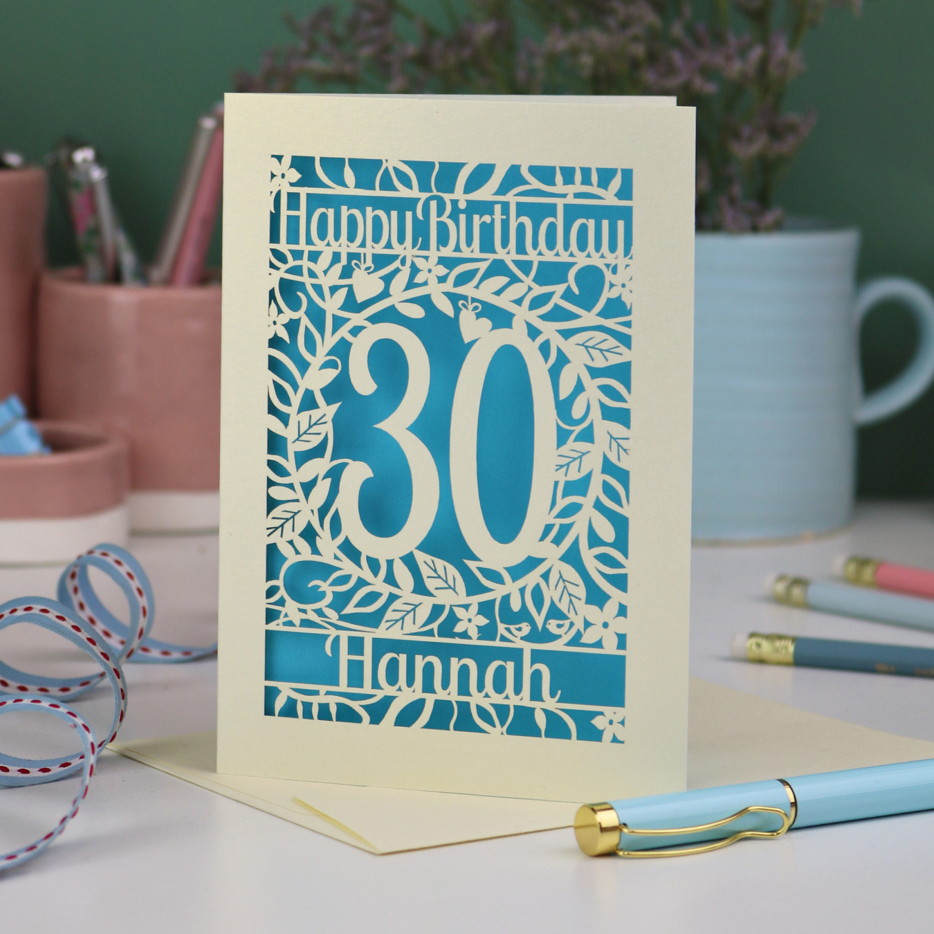 A laser cut floral 30th birthday card, personalised with the number 30 and the name Hannah, on a white desk with stationery items. 