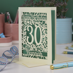 A laser cut floral 30th birthday card, personalised with the number 30 and the name Hannah, on a white desk with stationery items. 