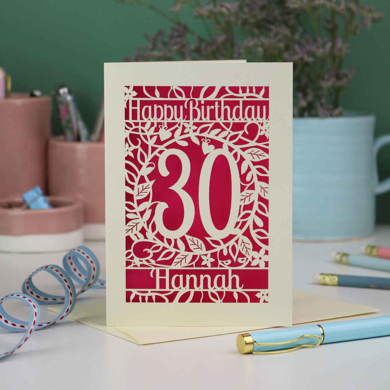 A paper cut floral 30th birthday card, personalised with the number 30 and the name Hannah, on a white desk with stationery items. 