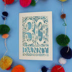Decorative paper cut out 30th birthday card with '30 Danny' on a blue background with colourful pom-poms.