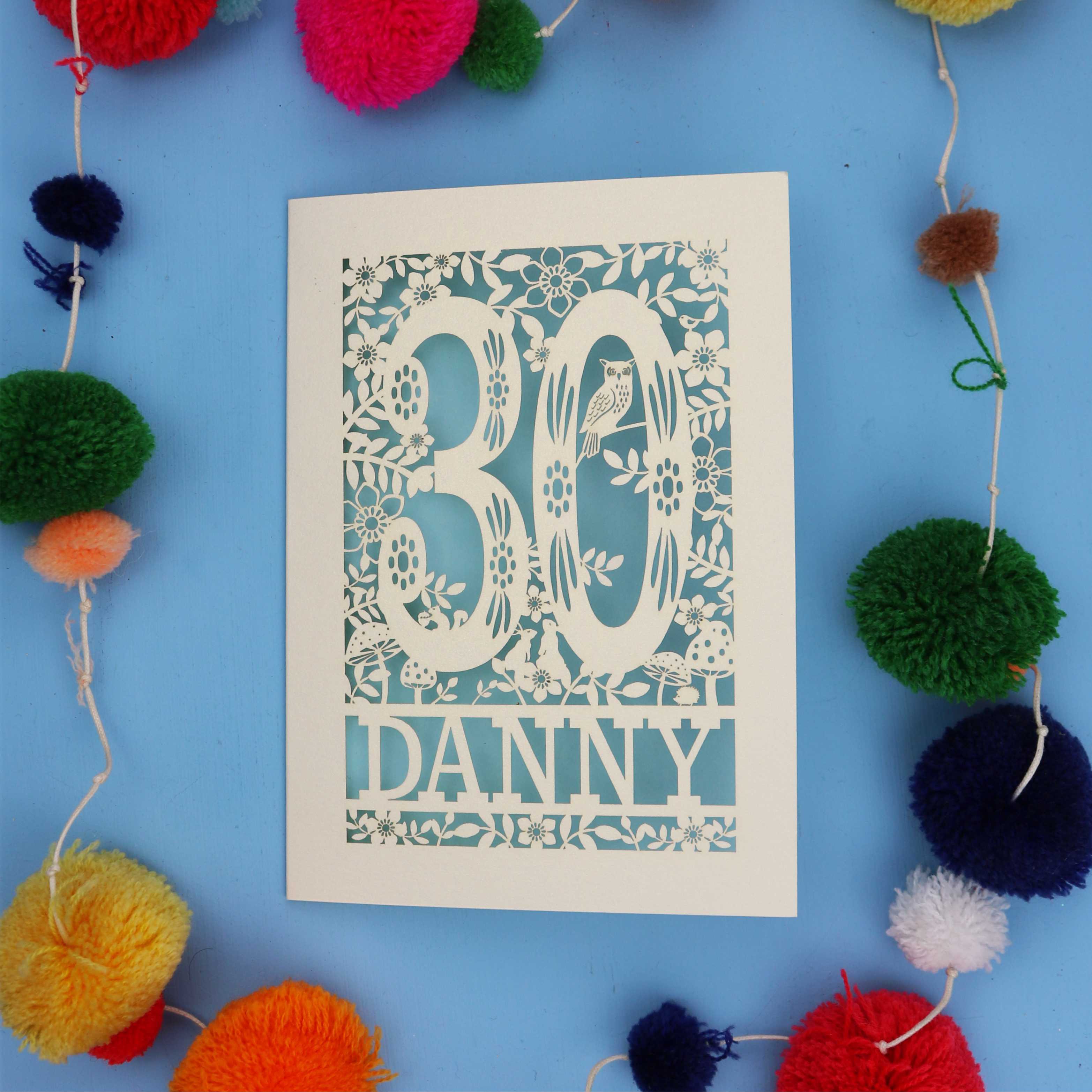Decorative paper cut out 30th birthday card with '30 Danny' on a blue background with colourful pom-poms.