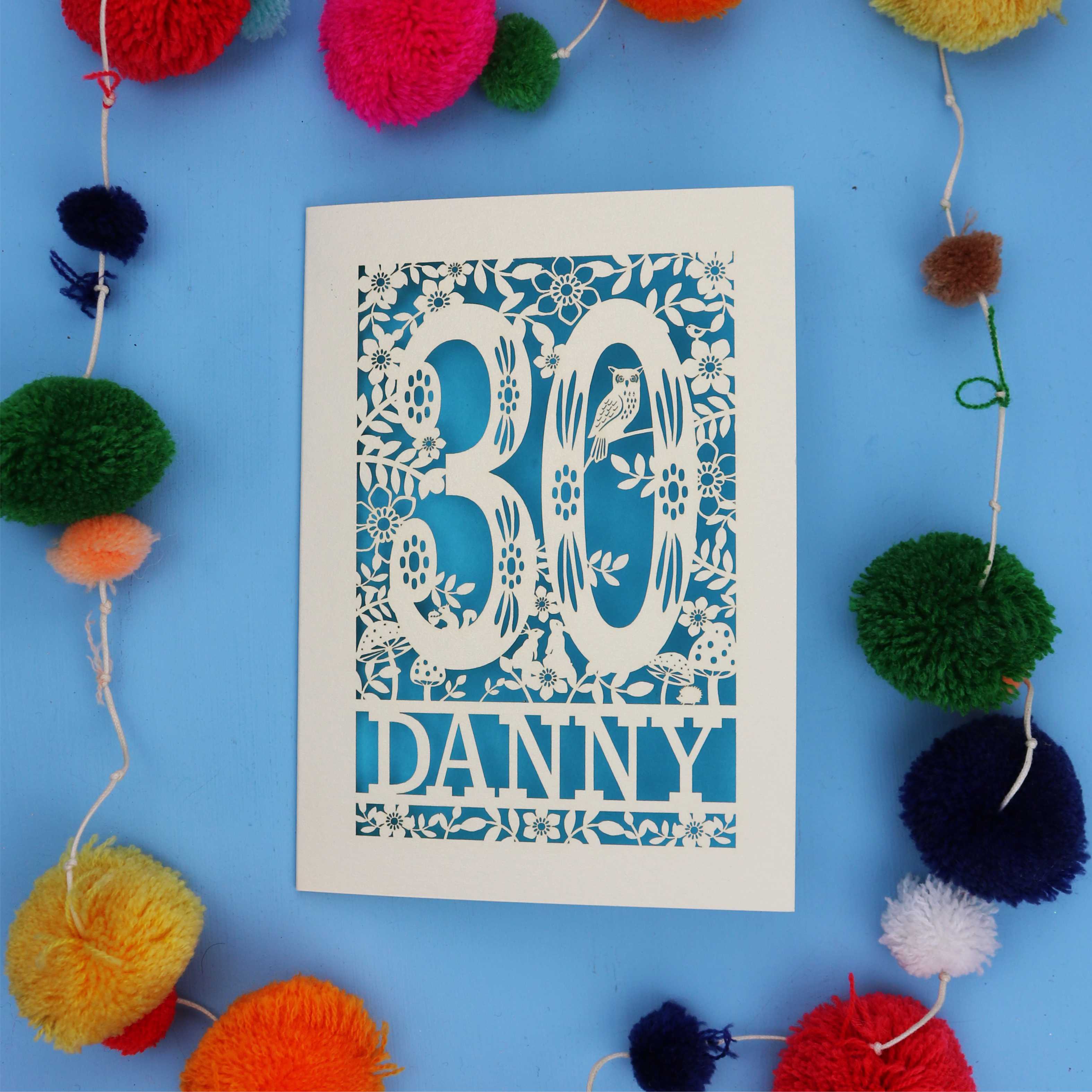 Decorative paper cut out 30th birthday card with '30 Danny' on a blue background with colourful pom-poms.