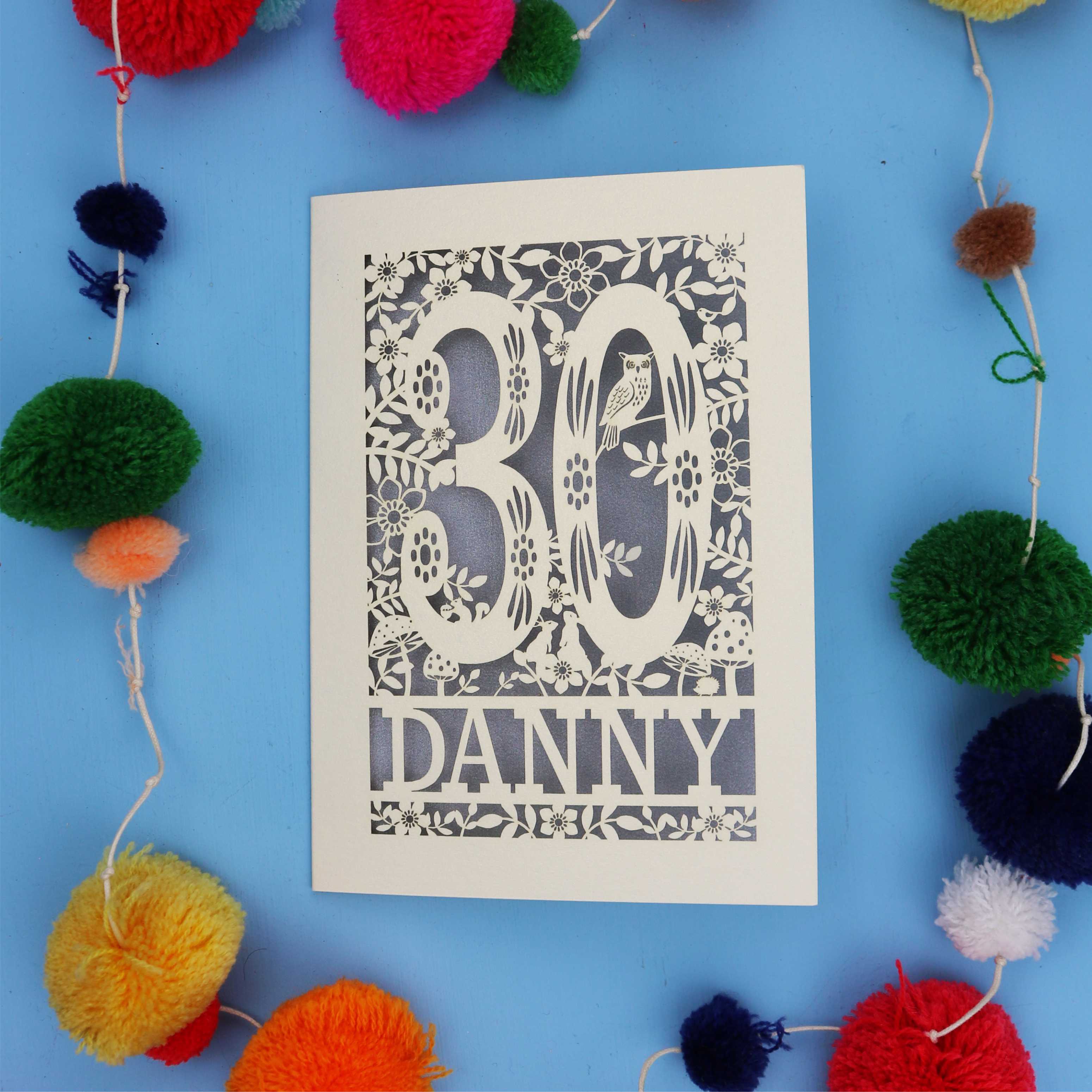 Decorative paper cut out 30th birthday card with '30 Danny' on a blue background with colourful pom-poms.