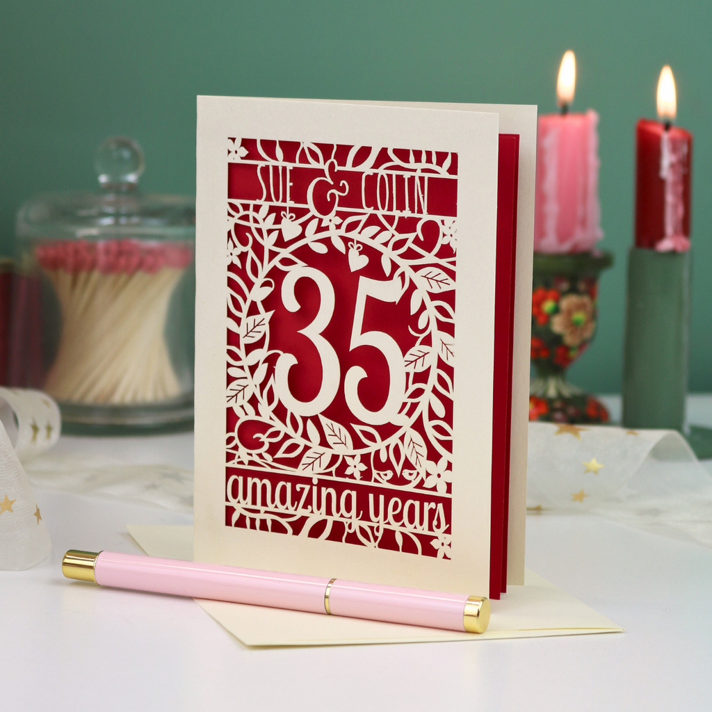 A personalised 35th anniversary laser-cut greeting card with a cream and red design, displayed on a white surface with a pink pen in the foreground and decorative candles in the background.