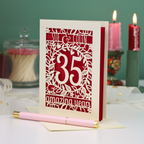 A personalised 35th anniversary laser-cut greeting card with a cream and red design, displayed on a white surface with a pink pen in the foreground and decorative candles in the background.