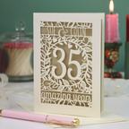A coral anniversary laser-cut greeting card with a cream and gold leaf design, displayed on a white surface with a pink pen in the foreground and decorative candles in the background.