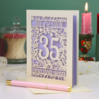 A 35th anniversary laser-cut greeting card with a cream and lilac design, displayed on a white surface with a pink pen in the foreground and decorative candles in the background.