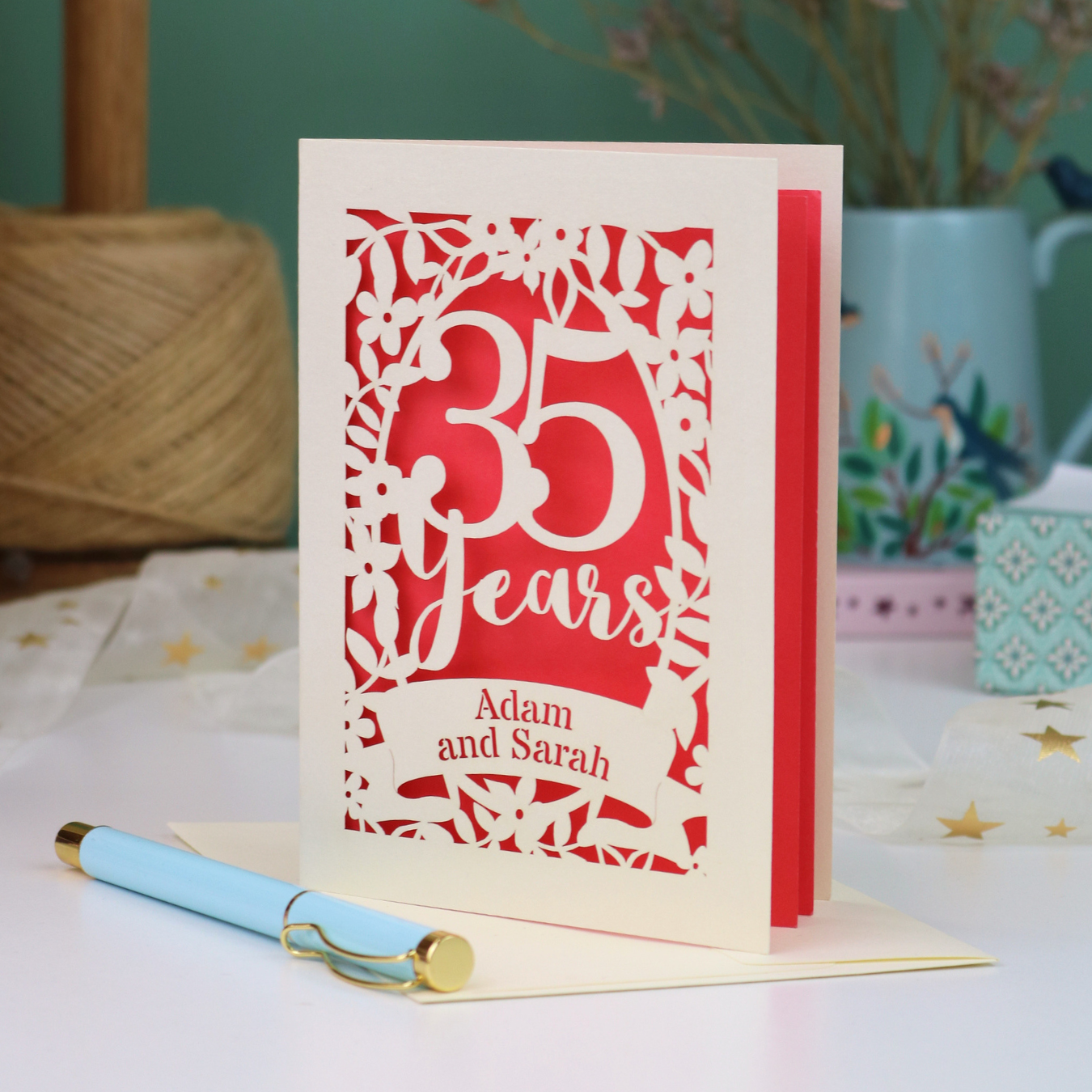 A personalized 35th anniversary card with laser-cut floral details and the text '35 Years Adam and Sarah', displayed on a table with a light blue pen in front. The background features a soft-focus teal wall, a vase of dried flowers, and a spool of thread.