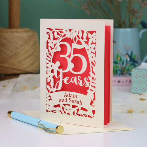 A personalized 35th anniversary card with laser-cut floral details and the text '35 Years Adam and Sarah', displayed on a table with a light blue pen in front. The background features a soft-focus teal wall, a vase of dried flowers, and a spool of thread.