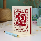 A personalised cut out 35th anniversary card with laser-cut floral details and the text '35 Years Adam and Sarah', displayed on a table with a light blue pen in front. The background features a soft-focus teal wall, a vase of dried flowers, and a spool of thread.
