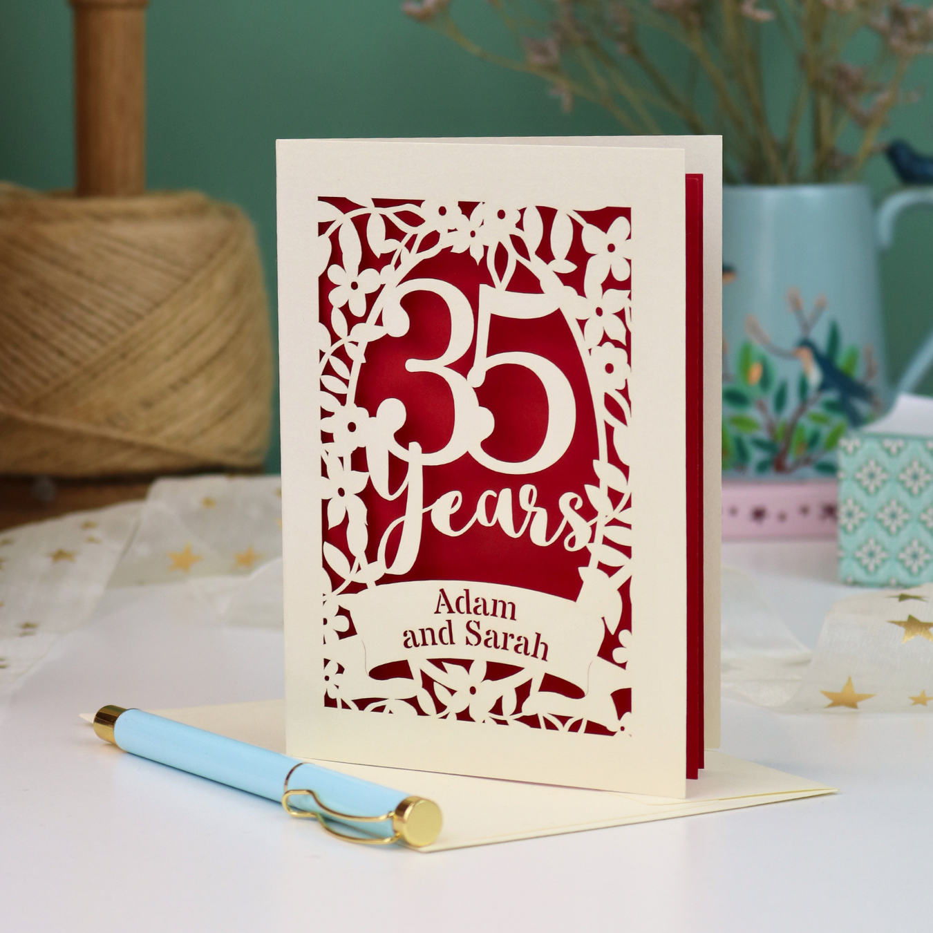 A personalised cut out 35th anniversary card with laser-cut floral details and the text '35 Years Adam and Sarah', displayed on a table with a light blue pen in front. The background features a soft-focus teal wall, a vase of dried flowers, and a spool of thread.