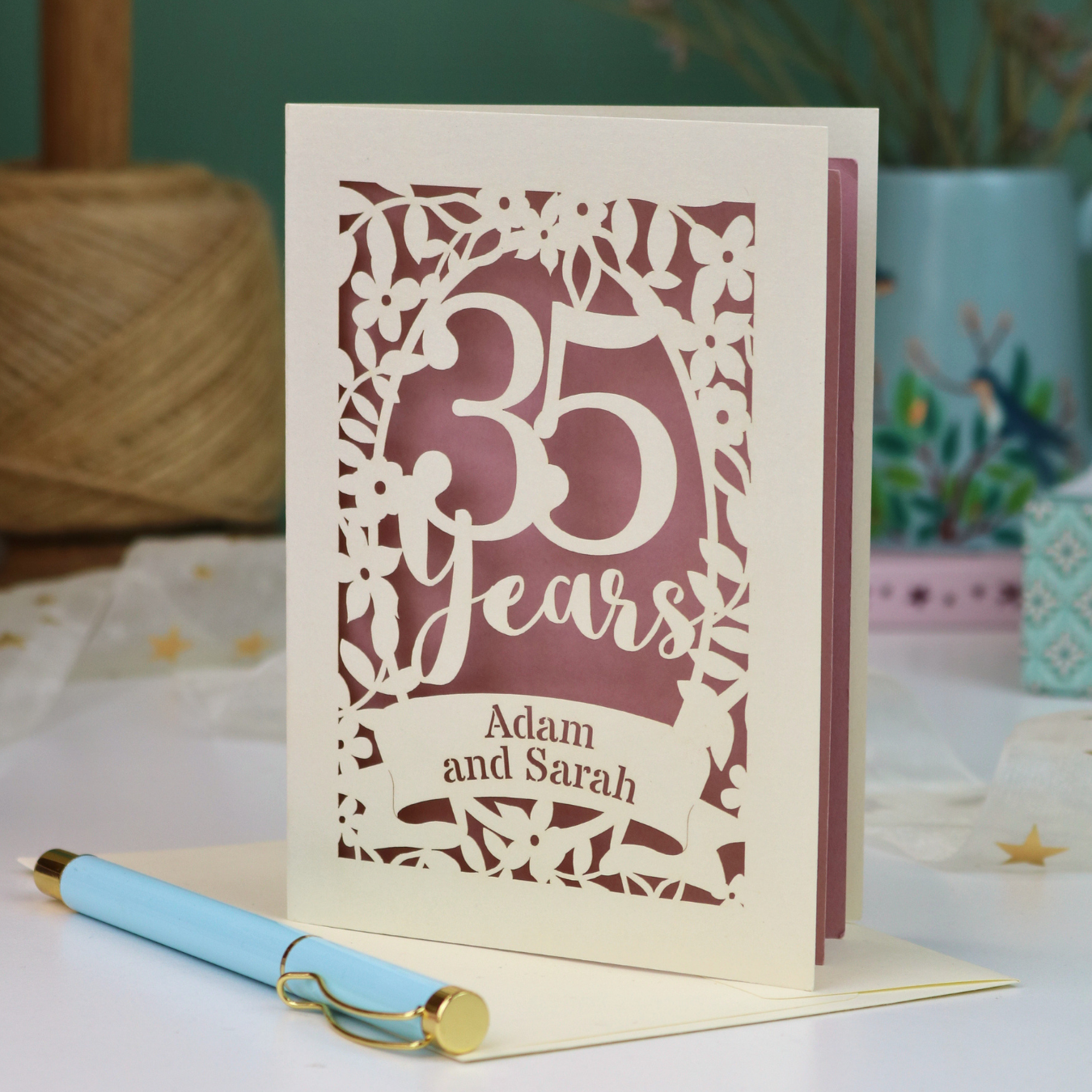A personalized coral anniversary card with laser-cut floral details and the text '35 Years Adam and Sarah', displayed on a table with a light blue pen in front. The background features a soft-focus teal wall, a vase of dried flowers, and a spool of thread.